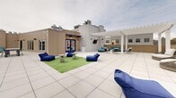 Located at 100 Hale Ave in White Plains, NY, this rooftop terrace offers a spacious and well-appointed outdoor retreat with striking views of the White Plains skyline. The terrace is finished with large-format light-colored tiles throughout, lending a clean, modern aesthetic to the entire space.

The layout is divided into several distinct seating and activity zones. Casual lounge areas feature plush navy blue bean bag chairs arranged on patches of artificial turf, accompanied by concrete side tables — ideal for relaxed gatherings. A dedicated seating section under a white pergola provides a shaded retreat, furnished with teal upholstered sofas and sleek metal chairs. An additional lounge area includes wicker-style lounge chairs, adding variety to the outdoor seating options.

A central fire pit with a round concrete surround serves as a natural focal point for evening gatherings, complemented by overhead string lights that create a warm, inviting atmosphere after dark. A greenery wall adds a vibrant natural contrast to the neutral building facade. The terrace also features an outdoor television mount and wall-mounted speakers, supporting both entertainment and events.

The open-sky exposure allows for abundant natural light, while the elevated position provides panoramic views of the surrounding White Plains cityscape, making this rooftop terrace a versatile and dynamic outdoor amenity.