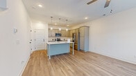 This two-story home is designed with a clean, contemporary aesthetic highlighted by light-toned hardwood-style flooring that runs consistently throughout both levels, creating a cohesive and bright atmosphere. The open-concept main floor features a generously sized living area with a ceiling fan, multiple windows allowing natural light to fill the space, and direct access to the entry. The kitchen is a functional centerpiece, equipped with gray shaker-style cabinetry, white countertops, a full suite of stainless steel appliances including a range, microwave, and French door refrigerator, and a central island with pendant lighting overhead — ideal for both cooking and casual dining. Upper-level bedrooms feature vaulted ceilings and ceiling fans, with windows providing natural light and neighborhood views. The primary suite includes a spacious walk-in closet outfitted with wire shelving systems for organized storage. The primary bathroom offers a wide double-sink vanity with gray cabinetry, white countertops, and a hallway leading to a window at the far end. A secondary bathroom includes a full soaking tub and shower combination. Throughout, the home maintains a neutral, move-in-ready palette.