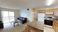 This single-floor space offers a functional and comfortable open-concept layout that seamlessly connects the living area and kitchen. The living room features plush carpeting and is furnished with a dark leather sofa and a low-profile coffee table, creating a cozy and inviting atmosphere. A sliding glass door fills the space with natural light and provides direct access to an outdoor balcony, enhancing the sense of openness.

The kitchen is positioned adjacent to the living area and is defined by a raised breakfast bar with a granite-style countertop, offering casual seating with wooden bar stools. Light wood cabinetry provides ample storage, while a full-size white refrigerator and additional appliances complete the setup. The kitchen's efficient layout makes it easy to navigate and well-suited for everyday use.

A hallway visible from the main living space leads to additional areas of the unit, including what appears to be a utility or storage closet. The textured ceiling runs throughout the space, and multiple HVAC vents ensure year-round climate control. Overall, the space presents a straightforward and practical layout with a neutral color palette that lends itself well to personalization.