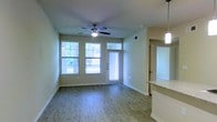 This single-floor space offers a clean, modern layout designed for comfortable everyday living. The main living area is bathed in natural light, courtesy of multiple large windows that frame views of lush greenery outside. A sleek ceiling fan with an integrated light fixture anchors the room, providing both airflow and ambient lighting. The flooring throughout is a warm, wood-style laminate that adds a welcoming and contemporary feel underfoot.

The living area flows naturally toward a front entry door flanked by a transom window, further enhancing the brightness of the space. Neutral, soft green-toned walls complement the light flooring and create a calm, versatile backdrop suited to a variety of interior styles.

Adjacent to the living area, a partial view of the kitchen reveals a pendant light fixture and a granite or stone-topped counter, hinting at a well-appointed cooking space. A corridor visible from the main area suggests additional rooms beyond, contributing to the functional layout of the floor plan.

White baseboards and clean trim work throughout add a polished finish to the interior. Overall, the space presents a bright, airy, and practical environment with thoughtful design details that balance style and function seamlessly.