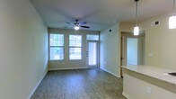 This single-floor space offers a clean, modern layout designed for comfortable everyday living. The main living area is bathed in natural light, courtesy of multiple large windows that frame views of lush greenery outside. A sleek ceiling fan with an integrated light fixture anchors the room, providing both airflow and ambient lighting. The flooring throughout is a warm, wood-style laminate that adds a welcoming and contemporary feel underfoot.

The living area flows naturally toward a front entry door flanked by a transom window, further enhancing the brightness of the space. Neutral, soft green-toned walls complement the light flooring and create a calm, versatile backdrop suited to a variety of interior styles.

Adjacent to the living area, a partial view of the kitchen reveals a pendant light fixture and a granite or stone-topped counter, hinting at a well-appointed cooking space. A corridor visible from the main area suggests additional rooms beyond, contributing to the functional layout of the floor plan.

White baseboards and clean trim work throughout add a polished finish to the interior. Overall, the space presents a bright, airy, and practical environment with thoughtful design details that balance style and function seamlessly.