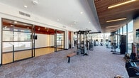 Located at 8555 East Raintree Drive in Scottsdale, Arizona, this well-appointed fitness center offers a generously sized, single-floor layout designed to support a full range of workout activities. The space is defined by its modern architectural character, featuring a striking combination of white walls, dark accent framing, slatted wood ceiling panels, and plush carpet flooring throughout the main exercise area.

Floor-to-ceiling glass windows line multiple walls, flooding the space with abundant natural light and providing open views of the surrounding Scottsdale landscape — including desert vegetation and neighboring buildings. A dedicated studio room, enclosed by sleek glass partition walls with black steel framing, features hardwood flooring and accent walls in deep red, making it ideal for group classes, yoga, or personal training sessions.

The main exercise floor is comprehensively equipped with a wide array of strength machines, including TKO cable and weight systems, multi-functional training rigs, and a full dumbbell rack. Cardio offerings include rowing machines, treadmills, ellipticals, and stationary bikes. An artistic mural accent wall adds a dynamic visual element to one section of the space, complementing the overall contemporary aesthetic. Ceiling fans and recessed lighting ensure comfort throughout. Large mirrors further enhance the sense of openness in the strength training area.