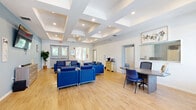 Located at 2124 Lepic Lane in Kissimmee, Florida, this single-floor space offers a well-appointed and inviting living area designed for comfort and functionality. The open layout is anchored by warm hardwood-style flooring that runs throughout, creating a bright and cohesive aesthetic.

The living room is the centerpiece of the space, furnished with multiple blue upholstered sofas and armchairs arranged to encourage conversation and ease of movement. A wall-mounted flat-screen television serves as a focal point, complemented by a dresser-style media console beneath it. A coffee station in the corner adds a practical, welcoming touch for visitors.

Architecturally, the coffered ceiling is a standout feature, adding depth and dimension to the interior while recessed lighting ensures even, warm illumination throughout. The light blue accent wall provides a calming contrast to the otherwise white and neutral palette, contributing to an overall atmosphere that feels open and serene.

Large windows with blinds line the walls, inviting ample natural light and offering views of the surrounding outdoor greenery. Double glass entry doors provide a grand yet accessible entrance. Additional interior doors lead to adjoining areas, suggesting further utility beyond the main living space. The overall ambiance is clean, modern, and thoughtfully designed to be both functional and comfortable.