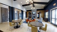 This contemporary communal lounge is a thoughtfully designed gathering space that blends modern aesthetics with comfortable functionality. The space immediately impresses with its soaring ceilings accented by bold dark exposed beams, creating a dramatic architectural framework that draws the eye upward. Elegant multi-bulb chandeliers are suspended throughout, casting warm, inviting light across the entire area.

A striking floor-to-ceiling stacked stone fireplace serves as the centerpiece of the main lounge area, flanked by a wall-mounted television and framed by warm wood paneling. The seating arrangement around the fireplace includes plush armchairs and sofas in rich orange and neutral tones, positioned atop a large area rug to define the relaxed conversation zone.

Toward the foreground, round dining tables paired with modern upholstered chairs in olive green and taupe tones provide casual seating for small groups. The light-toned tile flooring with a subtle herringbone pattern flows throughout, complementing the space's neutral and earthy color palette.

Dark accent walls contrast with bright white ceilings, while stone-clad columns add textural depth and visual interest. Adjacent areas, visible through open doorways, suggest additional rooms beyond, enhancing the sense of scale and connectivity throughout this well-appointed communal environment.