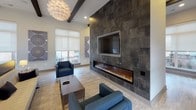 This single-floor communal lounge is designed with a contemporary aesthetic that balances comfort and sophistication. The most commanding architectural feature is a full-height dark stone tile accent wall housing a wide linear fireplace and a wall-mounted television, creating a striking focal point that anchors the main seating area. Light wood-style flooring runs throughout, providing a warm contrast to the bold stone surround.

The ceiling rises generously, accented by dark exposed wood beams that add architectural character and depth. Suspended from the ceiling are large, sculptural globe pendant lights that serve as decorative centerpieces while providing ambient illumination alongside recessed lighting.

The lounge is organized into distinct seating zones. A primary conversation area features a cream sofa, navy armchairs, and a low rectangular coffee table positioned near the fireplace. A secondary lounge area along the windowed perimeter offers paired gray accent chairs with small side tables, ideal for quiet seating.

Large windows fitted with roller shades line multiple walls, flooding the space with natural light and framing views of the surrounding outdoor areas. Double glass doors provide direct access to an exterior terrace. Decorative wall art and floor lamps complete the polished, welcoming atmosphere throughout.