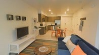 This single-floor unit offers a thoughtfully designed open-concept layout that seamlessly connects the living, dining, and kitchen areas into one cohesive, functional space. The living area is anchored by a plush navy blue sofa paired with a rustic round coffee table, all set atop a colorful striped area rug that adds vibrancy to the light-toned flooring. A white ladder shelf serves as a media and display unit, keeping the space organized yet stylish.

The kitchen is outfitted with warm wood-tone cabinetry in a two-tone arrangement, stainless steel appliances — including a refrigerator, range, and microwave — and a clean countertop workspace. Recessed ceiling lighting throughout ensures the space is evenly and warmly illuminated.

Between the kitchen and living area, a compact dining setup features a dark table surrounded by bold orange metal chairs, adding an energetic pop of color that ties together the room's accent tones. The entry area is neatly tucked near the dining space, with a clean white door and wall-mounted fixture completing the layout. The overall ambiance is modern and welcoming, with a cohesive design that balances practicality with contemporary style.