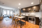This single-floor residence combines modern design with urban sophistication across a thoughtfully arranged layout. The open-concept kitchen and living area form the heart of the space, anchored by warm hardwood flooring that runs throughout. The kitchen is outfitted with dark, floor-to-ceiling cabinetry, a white subway tile backsplash, stainless steel appliances including a refrigerator, electric range, and built-in microwave, and a marble-topped island with seating for two — ideal for casual dining or entertaining. Recessed lighting illuminates the kitchen, while the adjoining living area benefits from expansive floor-to-ceiling windows that flood the space with natural light and offer views of the surrounding cityscape. The bedroom is a calm retreat featuring plush carpet, large windows with urban views, and well-considered proportions that comfortably accommodate a full suite of furniture. The bathroom is finished with classic white penny tile flooring, subway-tiled walk-in shower with glass enclosure, and dark vanity cabinetry. Dark-trimmed interior doors and neutral wall tones tie together the overall aesthetic, creating a cohesive, contemporary atmosphere throughout the residence.