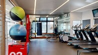 Located at 7555 E Warren Dr in Denver, CO, this fitness center offers a thoughtfully designed, single-floor exercise space that accommodates a full range of workout needs. The open exercise room is bathed in natural light from large windows along multiple walls, providing views of the surrounding outdoor environment and creating an energizing atmosphere for training.

The interior features a clean, modern aesthetic with sleek linear LED lighting embedded in white ceilings, durable rubber flooring, and full-length mirrors that enhance the sense of openness. Cardio equipment, including treadmills, ellipticals, and stationary bikes, is arranged along the windowed walls, each accompanied by mounted flat-screen televisions for entertainment during workouts.

The functional training zone is well-stocked, featuring an Octagon equipment rack loaded with a full dumbbell selection, medicine balls in various weights, plyo boxes, a heavy punching bag, TRX suspension trainers, and rowing machines. This variety supports strength training, conditioning, and dynamic movement exercises.

The entry area includes a water fountain station, key card access, and informational signage, offering a welcoming and organized arrival experience. Overall, the fitness center strikes a balance between compact efficiency and comprehensive equipment offerings, making it a capable facility for users of all fitness levels.