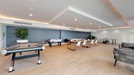 This single-floor amenity space is thoughtfully designed to serve both recreational and social functions, combining a vibrant game room with a relaxed lounge area in a seamless, open layout. Light hardwood-style flooring runs throughout, lending warmth and continuity to the contemporary aesthetic.

The game room section is equipped with a pool table, foosball table, air hockey table, ping pong table, and a shuffleboard table, catering to a wide range of recreational interests. A large communal wooden dining table with modern white and natural-toned chairs provides a casual gathering spot ideal for group activities or socializing. Recessed lighting and a tray ceiling with warm LED cove lighting add depth and sophistication to the space, while a muted blue-grey accent wall grounds the room visually.

Transitioning into the lounge area, floor-to-ceiling windows flood the space with natural light and offer views of a lush outdoor poolside area with tropical landscaping. Woven rattan lounge chairs with upholstered cushions are arranged for intimate conversation, complemented by minimalist side tables. A wall-mounted television and additional seating create a comfortable retreat. Potted tropical plants throughout add a refreshing, organic touch to the polished, modern atmosphere.