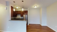 This single-floor space showcases a thoughtfully designed open-concept layout that seamlessly connects the kitchen and living area. The kitchen is a standout feature, outfitted with rich dark wood cabinetry that provides generous storage, granite countertops with subtle speckled detailing, and stainless steel appliances including a full-size refrigerator. A peninsula-style counter extends the workspace and offers the potential for casual seating, making it an inviting hub for cooking and entertaining. Pendant lighting hangs above the kitchen area, adding a stylish, focused illumination that complements the flush-mount ceiling fixtures throughout. Warm hardwood flooring flows continuously from the kitchen into the adjacent living area, creating a cohesive and inviting aesthetic. The living space is open and airy, with clean white walls and neutral tones that provide a versatile backdrop for any décor style. The well-maintained finishes and practical layout make this a functional and attractive space suited for a variety of everyday living needs.