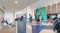 This fitness center in Spring, TX, is a thoughtfully designed, single-floor facility that combines high-end equipment with striking architectural details. The main workout area features soaring double-height ceilings and an expansive wall of floor-to-ceiling windows that frame serene waterfront views, flooding the space with natural light and creating an energizing atmosphere. A bold, large-scale teal mural depicting athletic imagery serves as a dramatic focal point, complemented by warm wood slat accents and vertical architectural columns throughout.

The strength training zone is well-equipped with a comprehensive rack of dumbbells, multiple weight benches, and a full lineup of Freemotion selectorized machines for targeted muscle training. Cardio equipment, including treadmills and upright cycles, is positioned directly along the window wall to take full advantage of the outdoor waterfront scenery. A dedicated functional training studio features a wood-slat ceiling with linear lighting, large wall mirrors, and an extensive collection of kettlebells, medicine balls, slam balls, and suspension training systems, providing ample space for dynamic and group-style workouts. The dual-zone layout and premium finishes give this fitness center a polished, resort-quality feel.