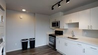 This single-floor space showcases a clean, modern aesthetic defined by a neutral palette and thoughtful design choices. The kitchen is a standout feature, outfitted with crisp white cabinetry equipped with sleek silver hardware, offering ample storage both above and below the countertops. Stainless steel appliances, including a gas range with oven and an over-the-range microwave, provide a polished, functional cooking environment. The countertops are finished in a light, neutral tone that complements the cabinetry, while a stainless steel sink with a contemporary faucet adds to the cohesive look. Overhead, a track lighting system with adjustable directional fixtures provides focused, customizable illumination throughout the kitchen area, supplemented by recessed lighting for an overall bright atmosphere. The dark hardwood-style flooring runs throughout the space, grounding the light tones of the walls and cabinetry with a warm contrast. The smooth, light-gray walls contribute to an open and airy feel. Overall, the space presents a well-coordinated, contemporary interior that balances style and practicality in a compact, efficient layout.