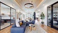 Located at 4500 S Monaco St in Denver, Colorado, this thoughtfully designed single-floor community clubhouse offers a dynamic blend of modern aesthetics and functional communal spaces. The expansive open-plan layout is anchored by a dramatic vaulted ceiling clad in warm wood planking, adorned with sculptural ring pendant lighting that serves as a striking focal point. Large arched and grid-pane black-framed windows flood the interior with natural light while framing views of the outdoor pool and surrounding greenery.

The main lounge area features plush sectional seating arranged over patterned area rugs atop light hardwood flooring, creating an inviting gathering space. A sunlit lounge nook near the oversized windows includes a linear fireplace set into a sleek shiplap-style accent wall, ideal for relaxed seating. Adjacent to the main lounge is a co-working or social lounge zone with built-in banquette seating, marble-top tables, and soft pendant lighting.

The fully appointed kitchen showcases an elongated waterfall-edge quartz island, sage green cabinetry, a textured tile backsplash, and stainless steel appliances — perfect for entertaining. Throughout the space, carefully curated artwork, indoor greenery, and warm wood tones contribute to a cohesive, upscale atmosphere that balances comfort with contemporary design.