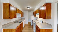 Located at 10450 SW McDonald St. in Tigard, Oregon, this two-bedroom, one-bathroom residence offers a functional and comfortable single-floor layout. The galley-style kitchen is a standout feature, lined on both sides with rich warm-toned wood cabinetry providing ample storage, complemented by light gray countertops, a double-basin sink, a white electric range, and a full-size refrigerator. Light hardwood-style flooring runs throughout the kitchen and flows into the combined kitchen-living area, where a ceiling fan and a large window invite natural light and outdoor views. The spacious living room continues the light flooring with clean white walls, creating an open and airy atmosphere. Both bedrooms feature plush carpeting and bright white walls; the primary bedroom offers a notably generous footprint with a window overlooking the surrounding neighborhood, while the second bedroom is cozy and well-proportioned. The bathroom is neatly appointed with a wood-toned vanity, light countertop, and a combination shower and tub. Textured ceilings are consistent throughout the residence. The clean, neutral palette and practical layout make this a move-in-ready home in a convenient Tigard location.