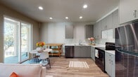 This two-floor space is designed with a clean, contemporary aesthetic throughout. The kitchen serves as a central highlight, outfitted with grey flat-front cabinetry, white countertops, and stainless steel appliances including a refrigerator, electric range, and dishwasher. Light wood-style vinyl flooring runs through the kitchen and open living/dining area, creating a warm, cohesive feel. Recessed lighting illuminates the space, and natural light enters through windows along the perimeter.

The dining area accommodates a dining set alongside the kitchen, with the layout offering an open, connected flow between cooking and living spaces. The bedrooms feature soft carpeting, neutral walls, and windows that allow natural light in, with one bedroom equipped with a wall-mounted mini-split HVAC unit for individual climate control.

Two bathrooms serve the unit: one features a sliding glass-door walk-in shower with grey vanity cabinetry, while the second offers a full tub-shower combination with matching grey cabinetry and a white countertop. An attached garage provides additional utility space, housing the water heater and a garage door opener, with a concrete floor and clean walls.