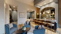 Located at 4909 France Ave S in Minneapolis, Minnesota, this single-floor communal lounge space offers a thoughtfully designed environment that blends sophistication with comfort. The open-plan layout accommodates a variety of social and leisure activities, anchored by several distinct yet interconnected zones.

At the heart of the space, a dramatic floor-to-ceiling dark marble feature wall frames a sleek linear fireplace, creating a striking focal point for the primary seating area. Plush gray upholstered armchairs and a sofa are arranged around a dark oval coffee table atop a textured area rug, inviting relaxed conversation. A secondary lounge nook, set against steel-blue accent walls with recessed paneling, offers a quieter retreat furnished with cream-toned sofas and swivel chairs around a marble-top table.

The wet bar area is a standout feature, showcasing an arched antiqued mirror backdrop, warm wood cabinetry with gold accents, pendant lighting, and a row of leather-upholstered bar stools. Large black-framed windows flood the space with natural light and provide views of the surrounding greenery. Wide-plank light hardwood flooring runs throughout, complementing the warm and neutral color palette. A billiards table rounds out the amenities, making this lounge a well-equipped and inviting gathering space.