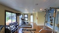This fitness center is a dedicated, single-floor exercise facility designed to accommodate a well-rounded workout experience. The space features a textured ceiling with recessed lighting that provides consistent, even illumination throughout. Large windows along one wall invite abundant natural light and offer views of the outdoor surroundings, creating an open and energizing atmosphere.

The layout is thoughtfully organized to house a variety of equipment. Cardio machines, including a treadmill and an elliptical trainer, are positioned centrally and make excellent use of the available floor space. A rack of dumbbells lines one wall, offering a wide range of free weights for strength and resistance training. On the opposite side, a weight bench and barbell rack provide additional options for targeted strength workouts.

The flooring is a durable, dark-toned surface well-suited for high-traffic exercise use. Neutral, light-toned walls contribute to the clean and functional aesthetic of the room. A clearly marked exit door with an adjacent fire extinguisher ensures safety compliance within the space. Overall, this fitness center delivers a practical, well-maintained environment with the essential equipment needed to support a comprehensive exercise routine.