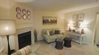 This single-floor space centers around a comfortable living area designed with a warm, inviting aesthetic. The room is anchored by a classic fireplace with a clean white mantle, serving as a natural focal point. Soft, neutral walls in a warm cream tone create a bright and airy backdrop throughout the space. Plush carpeting underfoot adds to the cozy feel of the room.

The living area is thoughtfully arranged with a sage green loveseat accented by decorative pillows in teal and coral tones, complemented by a striped accent chair. Two woven drum-style ottomans serve as a functional coffee table, adding a natural textural element to the space. Ambient lighting from a table lamp positioned on a slim console table along the wall provides a soft, warm glow, while framed artwork adds a refined decorative touch.

Wall décor includes a bold cityscape canvas painting above the seating area and a set of four circular medallion wall accents near the fireplace, adding visual interest and personality. The overall ambiance is relaxed yet polished, making the space feel both livable and stylish.