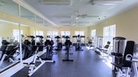 This fitness center offers a bright and welcoming environment designed to support a comprehensive workout experience. The space is generously sized, with a single open floor plan that accommodates a wide variety of exercise equipment, including treadmills, elliptical trainers, stationary bikes, and strength training machines arranged in an organized, easy-to-navigate layout. The flooring consists of durable, dark rubber matting, providing both safety and comfort during workouts. Soft yellow walls lend a warm, energizing ambiance to the room, while large windows along multiple walls flood the interior with abundant natural light and offer pleasant outdoor views. Ceiling fans are mounted overhead, ensuring comfortable airflow throughout the space. Full-length mirrors line portions of the walls, enhancing the sense of openness and allowing users to monitor their form during exercise. Recessed and flush-mounted lighting fixtures supplement the natural light, keeping the space well-lit at all times. The overall design emphasizes functionality and comfort, making this fitness center a practical and inviting facility for users of all fitness levels.
