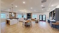 Located at 20 Franklin Ln in Manalapan, NJ, this thoughtfully designed single-floor space offers an expansive open-concept layout that seamlessly integrates a fully equipped kitchen with a generous living and gathering area. Light-toned hardwood flooring flows throughout, creating a warm and cohesive aesthetic that ties the entire space together.

The kitchen is appointed with natural wood cabinetry, a striking marble-veined quartz countertop, and a full suite of stainless steel appliances including a large refrigerator and dishwasher. A sizeable central island with bar seating serves as both a functional workspace and a social focal point, accented by elegant glass pendant lighting overhead.

The living area is anchored by a dramatic dark stone fireplace surround with a wall-mounted TV, flanked by a vertical wood slat accent panel that adds architectural texture. Multiple seating arrangements, including a plush sofa and rounded accent chairs, offer flexible gathering options. A built-in window bench with cushioned seating invites relaxed use alongside large windows that flood the space with natural light and provide views of the surrounding neighborhood. French doors lead to an exterior access point. Recessed lighting and a chandelier-style fixture provide layered illumination throughout, while a shuffleboard table and pool table add recreational appeal to this versatile community space.