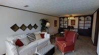 This single-floor home offers a comfortable and inviting layout designed for everyday living. The living room serves as the heart of the space, furnished with a generous white slipcovered sofa accented with decorative pillows and complemented by a bold red wingback armchair, creating a warm and casual atmosphere. A patterned ottoman adds a stylish focal point at the center of the seating arrangement. Decorative diamond-shaped accent pillows line the wall, adding a touch of personality to the space. A tall floor lamp and a lush potted plant bring warmth and a natural element to the corner of the room. The textured ceiling adds architectural character throughout. Toward the back of the living area, a set of arched built-in shelving units provides both display and storage space, flanking a ceiling fan with a light fixture that illuminates the transition into the dining area. The dining space features a dark metal-framed table and chairs, creating a functional and intimate setting for meals. Neutral-toned carpet flows throughout, tying the spaces together cohesively. Overall, the home presents a lived-in, welcoming ambiance with thoughtful decorative touches throughout.