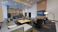 This single-floor communal space is designed to foster both relaxation and social interaction, combining a lounge area and a fully equipped kitchen within one open, airy environment. The room is defined by dramatically high ceilings that amplify the sense of openness, complemented by recessed lighting that provides consistent, even illumination throughout. The lounge area is anchored by a generous sectional sofa and modular seating in neutral tones, arranged around a solid wood coffee table, creating an inviting setting for casual gatherings. A large flat-screen television is mounted on a warm wood-paneled accent wall, flanked by built-in cabinetry that adds both function and aesthetic cohesion. Accent chairs in a deep charcoal tone provide additional seating options and a subtle contrast to the lighter furnishings. The carpeted flooring features a multi-tone pattern that grounds the space while adding visual texture. Toward the rear, the communal kitchen is equipped with professional-grade appliances, including ovens and ventilation hoods, alongside long communal dining tables with bench seating — ideal for group meals or events. The overall design balances a contemporary aesthetic with practical amenities, making it a versatile and welcoming shared environment.