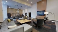 This single-floor communal space is designed to foster both relaxation and social interaction, combining a lounge area and a fully equipped kitchen within one open, airy environment. The room is defined by dramatically high ceilings that amplify the sense of openness, complemented by recessed lighting that provides consistent, even illumination throughout. The lounge area is anchored by a generous sectional sofa and modular seating in neutral tones, arranged around a solid wood coffee table, creating an inviting setting for casual gatherings. A large flat-screen television is mounted on a warm wood-paneled accent wall, flanked by built-in cabinetry that adds both function and aesthetic cohesion. Accent chairs in a deep charcoal tone provide additional seating options and a subtle contrast to the lighter furnishings. The carpeted flooring features a multi-tone pattern that grounds the space while adding visual texture. Toward the rear, the communal kitchen is equipped with professional-grade appliances, including ovens and ventilation hoods, alongside long communal dining tables with bench seating — ideal for group meals or events. The overall design balances a contemporary aesthetic with practical amenities, making it a versatile and welcoming shared environment.