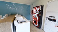 This single-floor laundry facility is designed for convenient, communal use. The space is anchored by a row of white top-load washing machines lined along one wall, providing ample capacity for multiple users. A wooden folding or sorting counter runs alongside the machines, adding practical surface space for laundry tasks. Dryers are also present within the room, rounding out the full wash-and-dry setup. The room features neutral tile flooring that is easy to maintain, and the walls are finished with clean white trim and crown molding. A standout design element is the blue accent wall adorned with decorative bubble graphics and a cursive "Laundry" script, lending the space a lighthearted, polished character. A bold red Coca-Cola vending machine positioned near the entrance adds a functional amenity for users waiting on their laundry. Overhead, a ventilation unit ensures proper air circulation throughout the room. Safety equipment, including a fire extinguisher, is visibly mounted on the wall. Overall, the facility strikes a balance between practicality and a well-kept, welcoming atmosphere.
