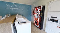 This single-floor laundry facility is designed for convenient, communal use. The space is anchored by a row of white top-load washing machines lined along one wall, providing ample capacity for multiple users. A wooden folding or sorting counter runs alongside the machines, adding practical surface space for laundry tasks. Dryers are also present within the room, rounding out the full wash-and-dry setup. The room features neutral tile flooring that is easy to maintain, and the walls are finished with clean white trim and crown molding. A standout design element is the blue accent wall adorned with decorative bubble graphics and a cursive "Laundry" script, lending the space a lighthearted, polished character. A bold red Coca-Cola vending machine positioned near the entrance adds a functional amenity for users waiting on their laundry. Overhead, a ventilation unit ensures proper air circulation throughout the room. Safety equipment, including a fire extinguisher, is visibly mounted on the wall. Overall, the facility strikes a balance between practicality and a well-kept, welcoming atmosphere.