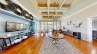 This well-appointed single-floor space offers a generously sized living room that immediately impresses with its striking architectural details and open, airy atmosphere. The centerpiece of the room is a beautifully crafted coffered ceiling with gold-toned insets and a decorative chandelier, lending a refined, classic elegance to the interior. Rich, warm hardwood flooring runs throughout, complementing the neutral and blue accent walls that add depth and character to the space.

Expansive floor-to-ceiling windows and multiple French doors line the walls, flooding the interior with an abundance of natural light and offering views of the surrounding outdoor areas, including what appears to be a pool and landscaped grounds. The openness created by the tall windows and high ceilings gives the space a bright, welcoming feel.

The living area is thoughtfully arranged into distinct functional zones. A central seating area features a round wood table atop a plush area rug, while leather armchairs and additional seating create comfortable conversation areas near the large windows. A dedicated workspace with a modern desk and upholstered chairs is positioned to take advantage of the natural light. A lean-to bookshelf and decorative wall accents add personality to the blue accent wall, completing the polished yet inviting ambiance of this versatile space.