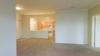 This single-floor space showcases an open-concept design that seamlessly connects the living area to the kitchen. The living space is finished with neutral-toned wall-to-wall carpeting and clean white walls accented by crown molding, lending a polished, cohesive feel to the interior.

The kitchen is accessed via a pass-through opening from the living area and features light oak cabinetry that provides ample storage. White appliances, including a range, built-in microwave, and refrigerator, complement the bright, functional layout. A peninsula with pendant lighting serves as both a workspace and a natural divider between the kitchen and living area, enhancing the open flow while defining each zone. The kitchen floor transitions to a hard surface, contrasting with the carpeted living space.

Overhead lighting in the kitchen, combined with the open pass-through design, allows light to carry throughout the shared space. The neutral color palette of warm beige and white tones creates a welcoming and versatile ambiance, well-suited for a variety of furnishing styles.