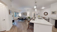 This single-floor residence is designed with an open-concept floor plan that seamlessly connects the kitchen, dining, and living areas into one cohesive, light-filled space. Wood-look flooring runs throughout the main living area, complementing the clean white walls and contemporary finishes.

The kitchen is equipped with stainless steel appliances, including a refrigerator, range, and microwave, alongside dark espresso cabinetry and a white countertop island that doubles as a breakfast bar with seating. Pendant lighting above the island adds a stylish focal point. The living area benefits from natural light through large windows, creating an open and airy atmosphere.

The primary bedroom is a comfortable retreat with neutral tones, a window, and an en-suite bathroom featuring a soaking tub, tile surround, dark wood vanity with a white countertop, and wood-look tile flooring. A second bedroom offers carpeted flooring and easy access to a second full bathroom, which includes a dark wood vanity and a large mirror. A dedicated in-unit laundry room houses a stacked washer and dryer alongside additional cabinetry for storage.