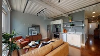 This single-floor loft showcases a compelling blend of industrial character and warm, eclectic style. The ceiling is a defining architectural feature throughout — white-painted corrugated metal panels with exposed beams, pipes, and track lighting lend an authentic industrial aesthetic to every room.

The open-concept living and kitchen area forms the heart of the space. The kitchen is outfitted with stainless steel appliances including a full-size refrigerator, range, microwave, and dishwasher, complemented by white cabinetry and open shelving. It flows seamlessly into the living area, which is anchored by a tufted tan leather sofa, a terracotta accent chair, and a richly patterned area rug. A wall of large windows floods the living room with abundant natural light and offers lush, tree-lined outdoor views, creating a bright and inviting atmosphere.

The bedroom continues the industrial-meets-warm design language, featuring the same distinctive ceiling, hardwood floors, an open closet, and vibrant accent decor. A corridor with polished concrete flooring connects the bedroom to the rest of the space.

The bathroom features a white vanity with ample cabinet storage, a large mirror with overhead lighting, and rich hardwood floors, providing a clean and functional finish to the unit.