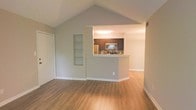 This single-story residence showcases a thoughtfully designed open-concept layout that seamlessly connects the living area to the kitchen. The living room is immediately distinguished by its dramatic vaulted ceiling, which rises to a sharp peak and creates a strong sense of vertical space and airiness. Neutral gray walls complement the warm-toned, hardwood-style flooring that runs continuously throughout the space, lending a cohesive and clean aesthetic.

A built-in recessed shelving unit is integrated into the living room wall, offering both functional storage and a decorative architectural detail. The kitchen is partially separated by a half-wall pass-through that features a breakfast bar ledge, providing both a visual boundary and a casual dining or workspace surface. The kitchen itself is equipped with stainless steel appliances, including a refrigerator and over-the-range microwave, alongside dark cabinetry that contrasts with the lighter tones of the surrounding space. Tile backsplash detailing adds texture to the kitchen's design.

White baseboards and trim throughout provide a crisp finishing touch, while a closet adjacent to the living area adds practical storage. The overall ambiance is bright, open, and move-in ready.
