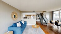 This sleek, urban apartment offers 1120 square feet of thoughtfully designed living space across two floors in the heart of downtown Austin. The open-concept main level showcases a well-appointed kitchen with stainless steel appliances, white cabinetry, and a unique copper-toned floor. The living area boasts floor-to-ceiling windows that flood the space with natural light and provide stunning city views.

The apartment features two bedrooms, including a spacious 230 square foot primary suite, and two full bathrooms. One bathroom showcases a modern design with a wood-look floor, while the other offers a more minimalist aesthetic. Both bedrooms are carpeted for comfort and include ample closet space.

A striking architectural element is the internal staircase with metal railings, leading to the upper level. This floor plan creates a sense of separation between living and sleeping areas. Throughout the apartment, neutral tones and contemporary furnishings create a sophisticated ambiance.

Additional features include recessed lighting, a dedicated closet space, and strategic placement of windows for optimal natural light. The property's location at 119 E 6th St places residents in the vibrant core of Austin, offering easy access to the city's renowned dining, entertainment, and cultural scenes.