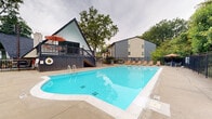 Located at 2814 De Mel Ave. in Louisville, Kentucky, this outdoor amenity space centers around a generously sized swimming pool with crystal-clear blue water. The pool area is thoughtfully designed for relaxation and recreation, featuring a wide surrounding deck finished in light concrete that provides ample space for movement and lounging. Multiple coral-toned lounge chairs are arranged along the perimeter, complemented by a shaded patio table set with a vibrant orange umbrella, offering a comfortable spot for outdoor seating. A secure black iron fence encloses the entire pool area, providing both safety and a defined boundary for the space. An elevated wooden deck is attached to a distinctive A-frame style building clad in white vertical board-and-batten siding with dark trim, accessible via a staircase and offering an elevated vantage point over the pool. Adjacent multi-story residential buildings with modern gray and neutral-toned exteriors are visible nearby. The space is richly framed by mature deciduous trees that provide a natural canopy and a sense of privacy, creating a serene, park-like atmosphere. The overall setting combines functional outdoor amenities with a welcoming, community-oriented ambiance.