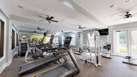 Located at 1600 Granby Street in Norfolk, Virginia, this fitness center offers a well-equipped, single-floor exercise space designed to support a full range of workout routines. The room features a neutral gray and white color palette accented by a bold yellow feature wall, creating an energetic yet focused atmosphere. Durable carpet flooring covers the entire space, providing comfort and stability underfoot during workouts.

The facility is outfitted with a comprehensive selection of equipment, including Matrix treadmills, elliptical machines, and cable-based strength systems from Paramount, along with a multi-station cable crossover unit. A dedicated free weight area along the yellow accent wall houses a full rack of dumbbells, a stability ball, and resistance accessories. Wall-to-wall mirrors on multiple sides visually expand the space and allow users to monitor their form throughout their workouts.

Ceiling fans are distributed throughout the room, ensuring consistent airflow and comfort during exercise. Natural light filters in through several windows with blinds, supplemented by recessed ceiling lighting. Flat-screen televisions are mounted on the walls for entertainment during cardio sessions. Glass-paneled double doors provide entry and exit points, contributing to an open, accessible feel. Overall, this fitness center delivers a practical and well-appointed environment for residents seeking convenient on-site exercise amenities.
