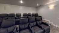 This single-level theatre is designed to deliver an immersive cinematic experience in a comfortable, well-appointed setting. The room is finished with cool grey walls and durable wood-look luxury vinyl flooring that runs throughout, lending a clean and modern aesthetic. Recessed ceiling lights and warm wall sconces provide layered ambient lighting, enhancing the room's inviting atmosphere.

Seating is arranged in two tiered rows of plush black leather reclining chairs, each equipped with built-in cup holders, ensuring every seat offers a clear sightline to the front of the room. The raised rear row further improves viewing angles, mirroring a true theatre layout. A large projection screen is mounted at the front of the room, framed by a wood-paneled accent wall and a curtain panel, adding a refined focal point to the space.

Practical details include a clearly marked emergency exit, a standard entry door with an adjacent dark wood door, wall-mounted speaker panels, and accessible control panels. The combination of thoughtful seating arrangements, quality finishes, and dedicated audiovisual infrastructure makes this theatre a well-equipped space suited for private screenings and group viewing events.