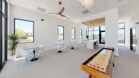 This single-floor communal amenity space is designed with a clean, contemporary aesthetic, characterized by crisp white walls, light-toned carpeting, and a thoughtful blend of open zones that serve different social functions. The layout is expansive and open, allowing natural light to flow freely throughout via large black-framed windows and full-height glass doors that connect the interior to an outdoor pool area.

The lounge zone features a plush sectional sofa, wall-mounted television, and a full-length wooden shuffleboard table, creating an engaging recreational focal point. Adjacent to this is a dining area furnished with multiple round tables and modern white chairs, ideal for group gatherings. Ceiling fans and a decorative pendant chandelier add functional and visual interest overhead.

A standout feature is the kitchen area, highlighted by a warm wood-plank ceiling, a generous white island with bar seating, stainless steel appliances, and a shimmering aqua tile backsplash beneath pendant lighting. A quieter seating nook with a sectional and ocean-themed artwork offers a more intimate retreat. Throughout, the design maintains a cohesive coastal-modern sensibility with warm wood accents contrasting against the predominantly white palette.