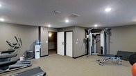 This fitness center is a compact yet fully functional workout space designed to meet a range of exercise needs. The room features neutral gray walls and a light carpet flooring, creating a clean and focused environment conducive to physical activity. Recessed ceiling lights, along with a central flush-mount fixture, provide bright, even illumination throughout the space. A multi-station cable machine and weight training equipment are positioned along one wall, offering users a comprehensive strength training setup. A treadmill is visible near the perimeter, catering to cardio exercise needs. A water cooler station is conveniently located in the corner, ensuring easy access to hydration during workouts. Two white panel doors lead to adjacent rooms, likely changing or utility spaces, adding to the overall functionality of the facility. The layout is open and uncluttered, allowing for ease of movement between equipment. The space presents a practical and welcoming environment suitable for residents or guests looking to maintain an active lifestyle without leaving the building.