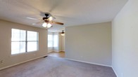This single-story space offers a comfortable and functional layout across its open living areas. The main living room is generously proportioned, finished with wall-to-wall carpeting in a neutral tone that complements the soft beige walls and white baseboards. A ceiling fan with a light fixture provides both ventilation and ambient lighting, contributing to a practical and understated aesthetic. A standard window along one wall allows natural light to filter into the space.

The adjoining room transitions to hardwood-style flooring, offering a visual distinction between the two areas. This secondary space features a notable bay window that curves outward, inviting an abundance of natural light and adding architectural interest to the otherwise straightforward layout. A second ceiling fan mirrors the one in the main living area, maintaining a consistent design throughout.

The textured ceilings and neutral color palette throughout create a calm, move-in ready environment. The open sightlines between the two rooms enhance the overall sense of space, making the layout feel connected and airy. The combination of carpet and hard flooring provides versatility suited to a range of furnishing styles.