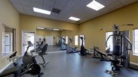 This fitness center offers a practical and well-organized workout environment designed to support a range of exercise routines. The space features warm yellow walls and a drop-tile ceiling fitted with recessed fluorescent lighting panels that ensure consistent, even illumination throughout. A large floor-to-ceiling mirror spans one wall, visually expanding the room and allowing users to monitor their form during exercise. The durable, smooth concrete flooring provides a sturdy surface suited for fitness equipment and high-traffic use. Natural light filters in through multiple windows, adding brightness and an open feel to the interior. The equipment layout includes a variety of cardio machines such as stationary bikes, elliptical trainers, and a recumbent bike, alongside strength and resistance equipment including a cable machine and weight station. The machines are arranged with ample space between them, allowing for comfortable movement and safe use. The space also benefits from a visible entry door that connects to an adjacent area, hinting at its role within a larger facility. Overall, this fitness center provides a clean, functional, and accessible environment for a comprehensive workout experience.