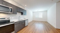 Located at 5300 E Washington St in Phoenix, AZ, this single-floor unit offers a thoughtfully designed layout that maximizes its compact footprint. The open-concept design seamlessly connects the kitchen and living area, creating a functional and cohesive living space. Light-toned hardwood-style flooring runs throughout, contributing to a clean, contemporary aesthetic that is consistent across every room.

The kitchen is well-equipped with a full suite of stainless steel appliances, including a refrigerator, electric range, and over-the-range microwave. Dark charcoal cabinetry with brushed silver hardware provides ample storage, while white countertops offer a crisp contrast. A stainless steel sink with a modern pull-down faucet and an integrated dishwasher complete the practical setup.

The living area is bright and open, with a window fitted with horizontal blinds that allows natural light to fill the space. White walls and recessed lighting enhance the airy atmosphere. A hallway connects to the bathroom, which features a full bathtub with shower, a vanity, and consistent wood-style flooring. The unit also includes in-unit laundry, adding notable convenience to this efficiently designed space.