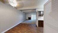 This single-floor residence offers a well-organized layout with a blend of practical design elements and comfortable living spaces. The open-concept living and dining area is defined by a textured popcorn ceiling with distinctive exposed beams, rich hardwood-style flooring, and neutral-toned walls that create a warm, inviting atmosphere. A classic brick fireplace serves as a focal point in the carpeted living room, which also features a sliding glass door with vertical blinds that allows natural light to flow in and provides access to an outdoor area. The kitchen is efficiently designed with dark cabinetry, a white countertop, and a range, partially separated from the main living area by louvered bifold doors that add a retro character to the space. The bedrooms are carpeted and feature the same exposed beam ceiling detail, with windows that bring in natural light. The bathrooms are equipped with accessibility features, including grab bars in both the tiled walk-in shower — which features a mosaic tile floor — and the separate bathtub with overhead shower, both finished in neutral cream tiling. Throughout the space, the consistent architectural details of exposed beams and textured ceilings tie the rooms together into a cohesive and functional home.