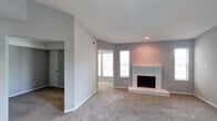 This single-story space offers an open and airy living area defined by vaulted ceilings that enhance the sense of volume and openness. The room is finished with neutral gray walls and wall-to-wall carpet, creating a clean, cohesive aesthetic throughout. A white brick fireplace serves as a prominent focal point, featuring a raised hearth and a display shelf above the firebox, adding both architectural character and functional warmth to the space. Recessed lighting supplements the natural light that filters in through multiple windows, keeping the interior bright and welcoming. The open layout connects seamlessly to an adjacent area, visible through a wide entryway, which provides flexibility for various uses. A hallway leading to additional rooms is accessible from the main living space, with a door visible at the end suggesting further private areas beyond. The textured ceiling, crisp white baseboards, and consistent neutral color palette contribute to a tidy, move-in-ready presentation. Overall, the space balances everyday functionality with comfortable, understated design elements suited to a range of living arrangements.