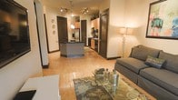 This single-floor residence offers a well-designed open-concept layout that connects the living and kitchen areas in a cohesive, functional flow. The living area is anchored by warm hardwood flooring, a plush sofa, and a glass-top coffee table resting on a decorative area rug, creating an inviting and comfortable space. A tall accent lamp adds warm ambient lighting that complements the natural tones throughout.

The kitchen is a standout feature, equipped with stainless steel appliances including a refrigerator and range, dark countertops, and light wood cabinetry that extends toward the ceiling. A central kitchen island with a dark countertop provides additional prep space and serves as a natural dividing element between the kitchen and living area. Pendant lighting and a track lighting system illuminate the space effectively, enhancing the modern aesthetic.

Dark-finished doors and bold baseboards throughout create a striking contrast against the warm, neutral walls, adding a sense of architectural definition. A louvered door adds a classic touch, contributing to the layered design details of the home. Overall, the space presents a balanced combination of modern finishes and warm, welcoming tones that make it both practical and visually appealing.