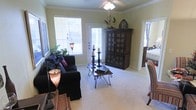 This single-floor space centers around a well-appointed living room that combines classic architectural details with a warm, inviting atmosphere. Crown molding lines the ceiling perimeter, adding a refined touch, while a ceiling fan with a light fixture provides both comfort and illumination. The room is finished with plush wall-to-wall carpeting in a neutral tone, contributing to a cozy and cohesive feel throughout.

Natural light is a defining characteristic of the living area, with multiple windows fitted with horizontal blinds and a glass-panel exterior door that allows daylight to fill the space. The pale yellow walls enhance the brightness, creating an airy environment.

The living room is thoughtfully laid out to accommodate comfortable seating and functional furnishings, including space for a large freestanding armoire with glass-front cabinet doors, which serves as a prominent focal point. A doorway visible from the main area suggests seamless connectivity to additional rooms within the home. Overall, the space presents a traditionally styled, light-filled interior with classic detailing throughout.