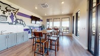 This single-floor communal space is designed for social gathering and relaxation, combining an energetic aesthetic with functional amenities. The interior is immediately eye-catching, featuring bold, professionally executed graffiti murals that span entire walls — including a vivid "Chill Up" mural in pink and teal tones and a striking purple-and-gray mascot-inspired design. These artistic elements give the space a dynamic, youthful character that sets it apart.

The main gathering area is outfitted with multiple bar-height tables and wooden stools, encouraging casual seating arrangements suited for group hangouts or social events. Large black-framed windows flood the interior with natural light and provide open views toward the adjacent outdoor terrace. Durable wood-look flooring runs throughout the space, complementing the warm, neutral wall tones.

A built-in kitchenette features sage-green cabinetry, a double stainless steel sink with a white subway tile backsplash, and ample counter space — ideal for hosting or refreshments. Wall-mounted flat-screen TVs are positioned for clear sightlines from the seating areas.

The outdoor terrace is a standout feature, offering a covered bar-height counter along a glass-railed balcony with sweeping panoramic views of surrounding mountains and the cityscape below. A ceiling fan and mounted TV make the terrace a comfortable outdoor retreat in nearly any weather. The overall ambiance is lively, modern, and inviting.