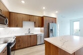 This single-floor residence offers a thoughtfully designed layout encompassing two bedrooms, two bathrooms, a combined kitchen and living area, a hallway, and two closets. The interior design reflects a contemporary aesthetic, combining warm walnut-toned cabinetry with crisp white surfaces throughout.

The kitchen serves as a central highlight, featuring floor-to-ceiling wood cabinetry, a striking Calacatta quartz backsplash and countertops, stainless steel appliances including a French door refrigerator, dishwasher, range, and microwave, alongside a generous island that provides additional prep space and storage. Light wood-tone flooring flows throughout the open-concept kitchen and living area, enhancing the bright, airy atmosphere created by recessed lighting.

The spacious primary bedroom offers generous proportions and direct access to a dedicated closet with built-in hanging rods. The en-suite bathroom features a frameless glass walk-in shower with full marble-style tile surround, a wood-vanity with quartz countertop, and matte black fixtures. The second bathroom mirrors the refined aesthetic with a soaking tub, marble-style tiled surround, and coordinating fixtures. Both bedrooms display light hardwood flooring and clean white walls, creating a neutral, versatile canvas.