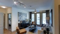 This single-floor residence offers a thoughtfully designed living area that combines contemporary style with functional comfort. The main living space is anchored by a striking large-scale abstract artwork in muted blue and gray tones, mounted above a sleek, dark credenza that serves as a media console and display surface. Track lighting runs along the ceiling, providing adjustable, directional illumination that complements the ceiling fan with an integrated light fixture positioned at the room's center.

The space benefits from generous natural light, courtesy of tall floor-to-ceiling windows and glass doors dressed with dark curtain panels and horizontal blinds. These openings offer views of the surrounding exterior and lend an open, airy quality to the interior. Light-toned walls and light hardwood-style flooring enhance the brightness throughout.

The seating arrangement features a neutral sofa, cognac leather accent chairs with a modern frame, and a dark rectangular coffee table, all anchored by a patterned area rug. The overall ambiance is modern and sophisticated, with a carefully curated mix of textures and tones that create an inviting yet polished atmosphere.